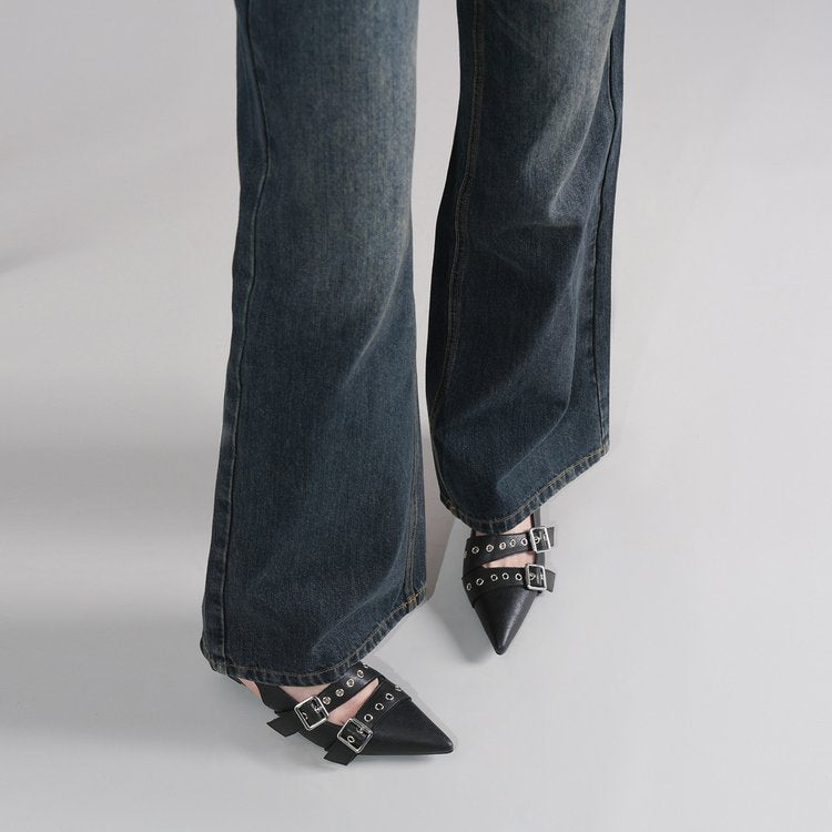 A close-up of a person's legs wearing dark blue wide-leg jeans and black pointed-toe flats with silver buckle straps, shown against a light gray studio background.