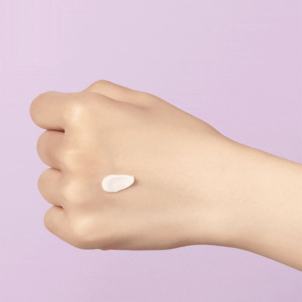 A close-up of a person's hand with a small dollop of white cream or lotion on the back of the hand, set against a solid light purple background.