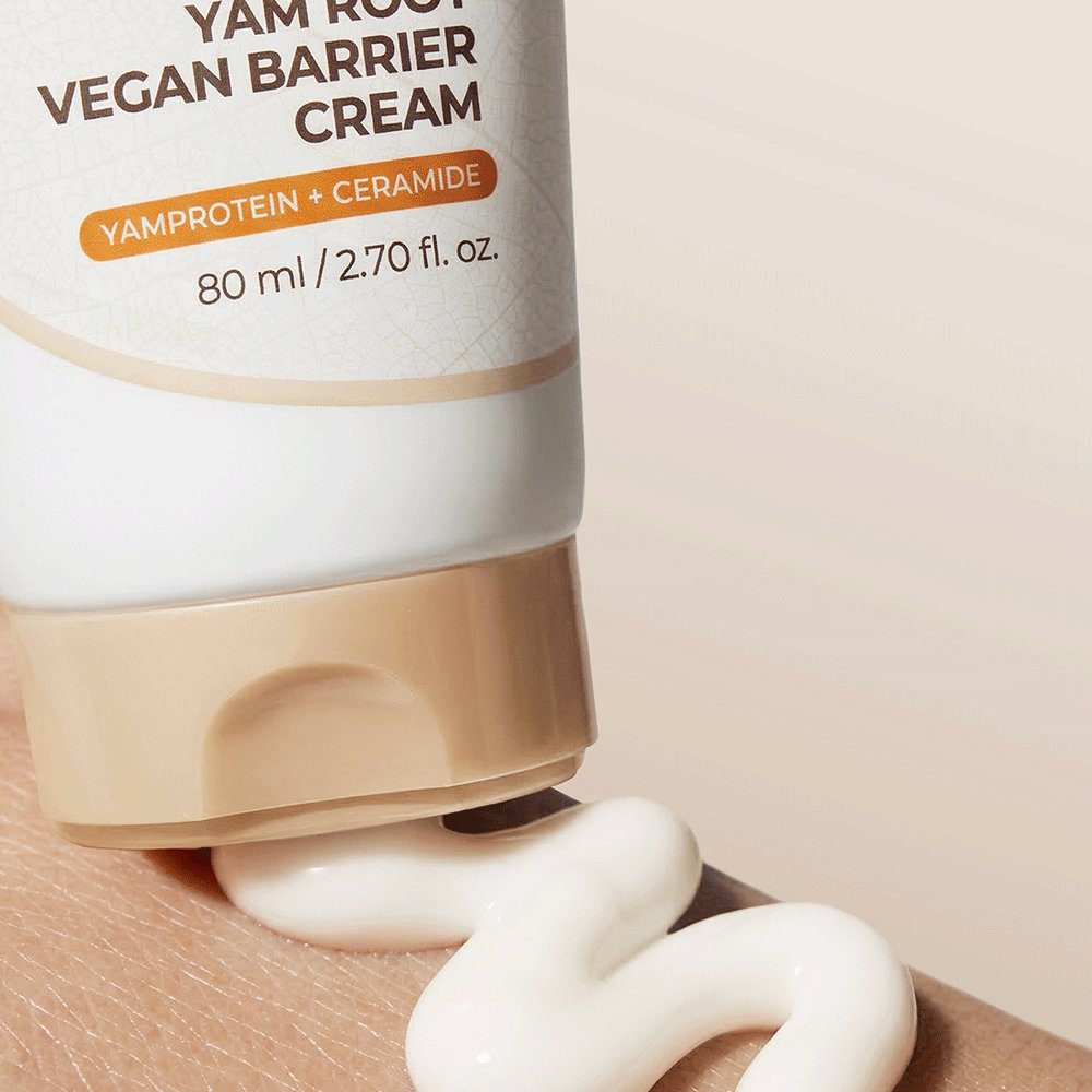 A close-up of a YAM ROOT VEGAN BARRIER CREAM tube dispensing thick white cream onto a beige surface. The tube is white with a beige cap and features orange and brown text.
