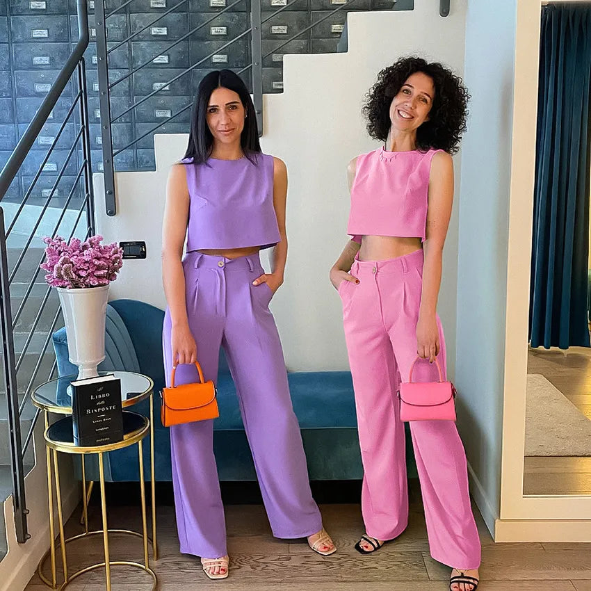 Two women in matching outfits with colorful handbags standing in a modern interior setting.