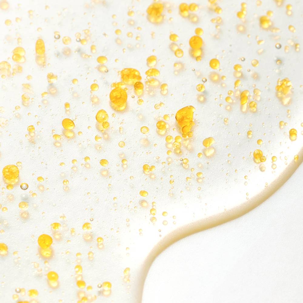 A close-up of a clear, glossy liquid cosmetic or skincare product with scattered translucent golden-yellow droplets on a white surface.