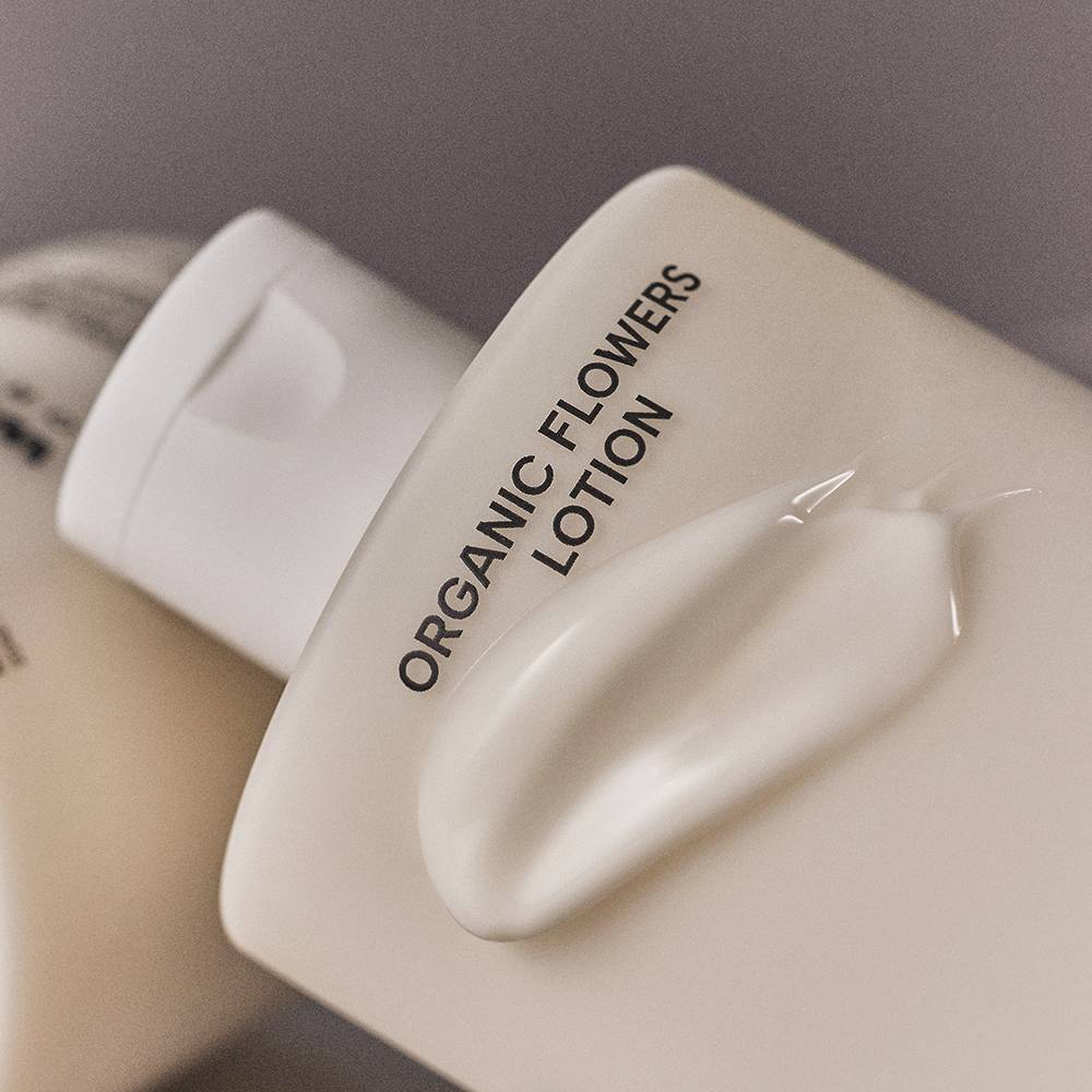 A close-up of a white cosmetic bottle labeled 'ORGANIC FLOWERS LOTION' with a glossy smear of white lotion on the front, set against a soft gray background.