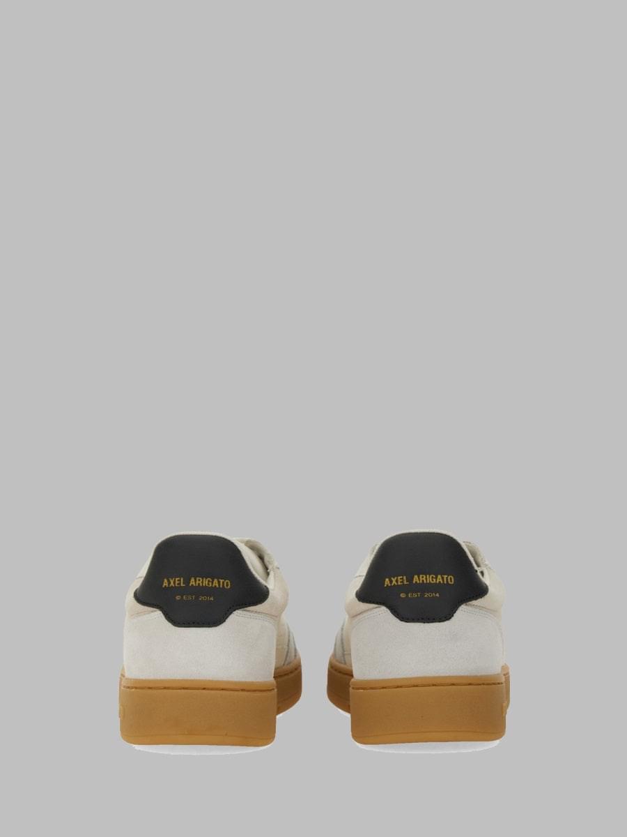 A pair of low-top sneakers shown from the back, featuring off-white suede and leather panels, a black heel tab with gold 'AXEL ARMANIATO' branding, and tan gum soles, set against a solid gray background.