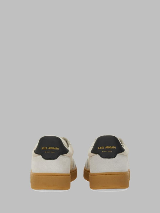 A pair of low-top sneakers shown from the back, featuring off-white suede and leather panels, a black heel tab with gold 'AXEL ARMANIATO' branding, and tan gum soles, set against a solid gray background.