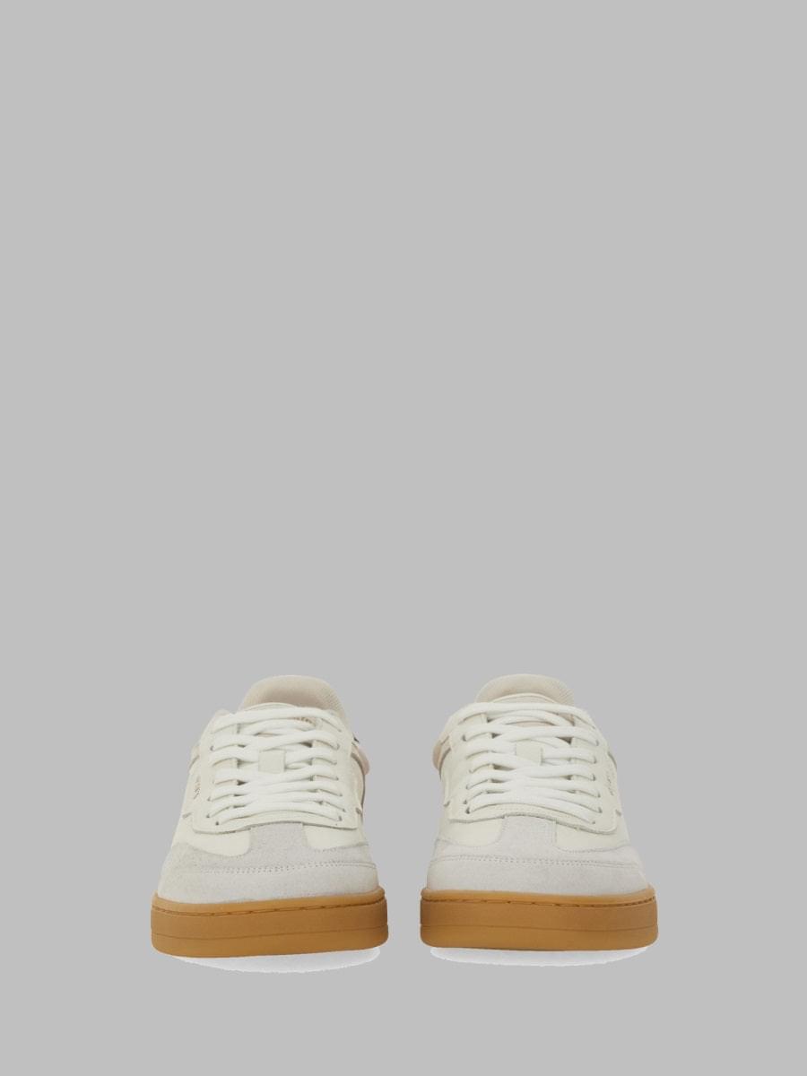 A pair of low-top sneakers in off-white leather and suede with white laces and gum rubber soles, shown from the front against a solid gray background.
