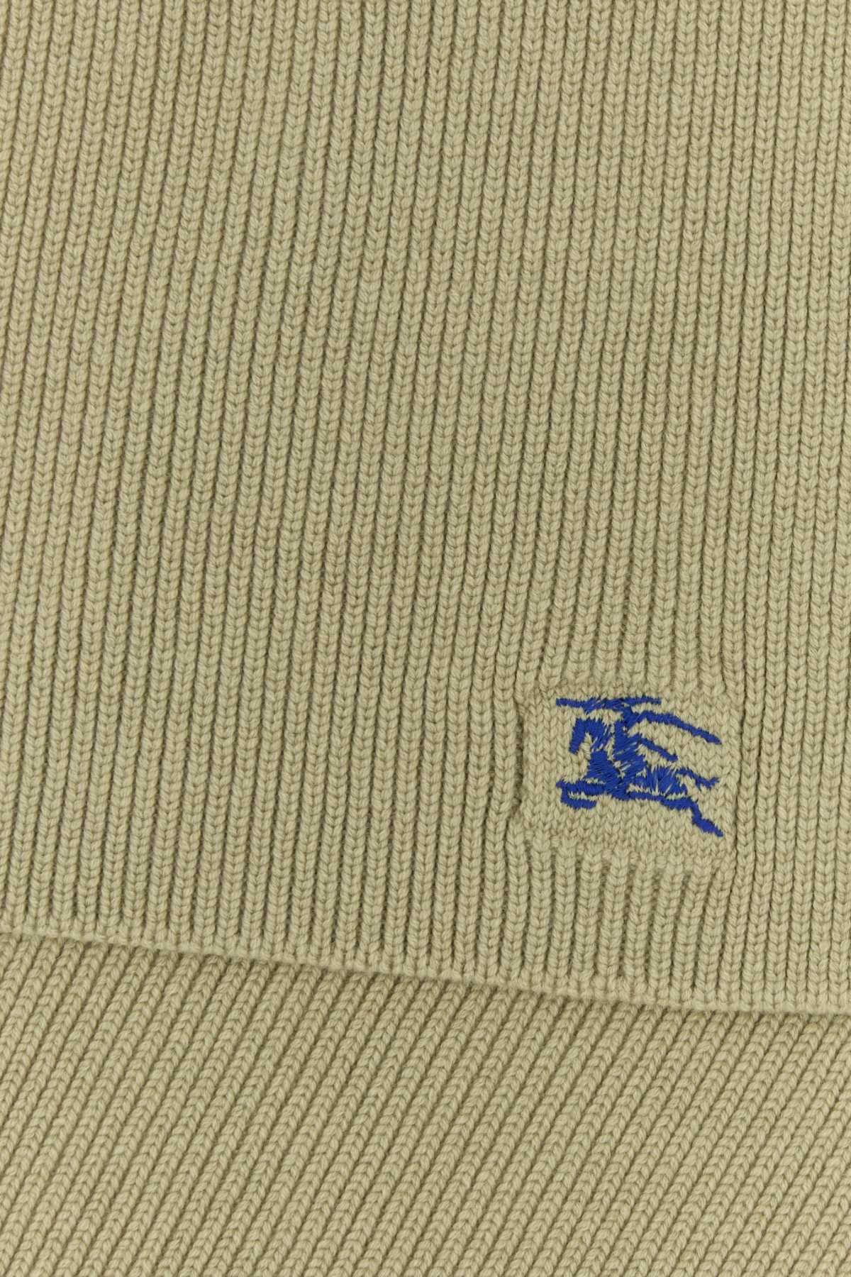 Close-up of a ribbed knit fabric in a light khaki-beige color, featuring a small embroidered blue logo patch on the lower right.