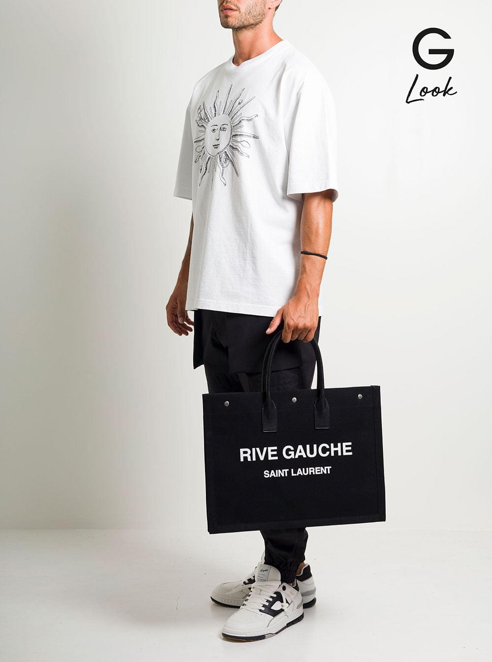 A person wearing a casual outfit while holding a black Saint Laurent Rive Gauche canvas tote bag, set against a clean, minimalist white studio background.
