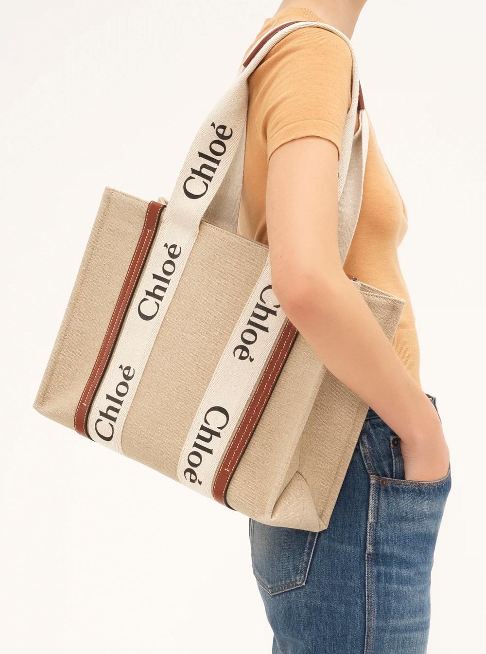 A woman stands against a minimalist, neutral background, elegantly carrying the Chloé 'Woody' medium tote bag in beige linen with brown leather trim and prominent logo detailing.