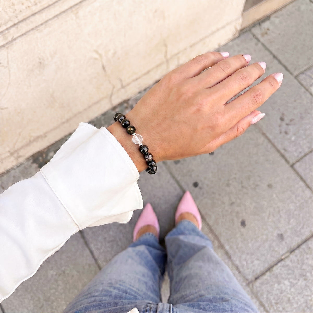 A person wearing a black beaded bracelet with a small clear crystal accent, shown on their wrist in an outdoor setting with gray pavement and a beige wall in the background. The Hypersthene Millionaire Manifestation Bracelet - Rare Egyptian Wealth Stone