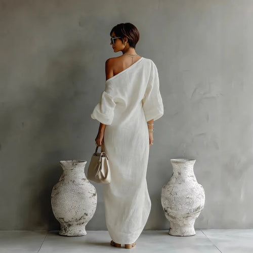 Woman in a white dress standing between two large vases against a gray wall.