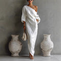 Woman in a white one-shoulder dress standing between two large vases against a gray wall.