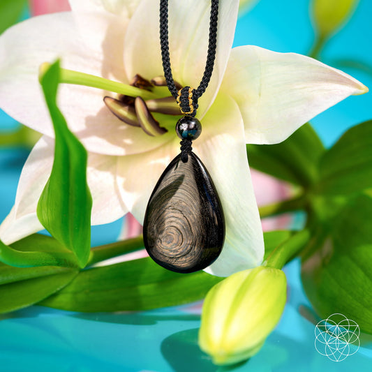 Black teardrop pendant on a necklace with a white flower and green leaves in the background. The ﻿Hypersthene Millionaire Manifestation Pendant. Rare Egyptian Wealth Stone.