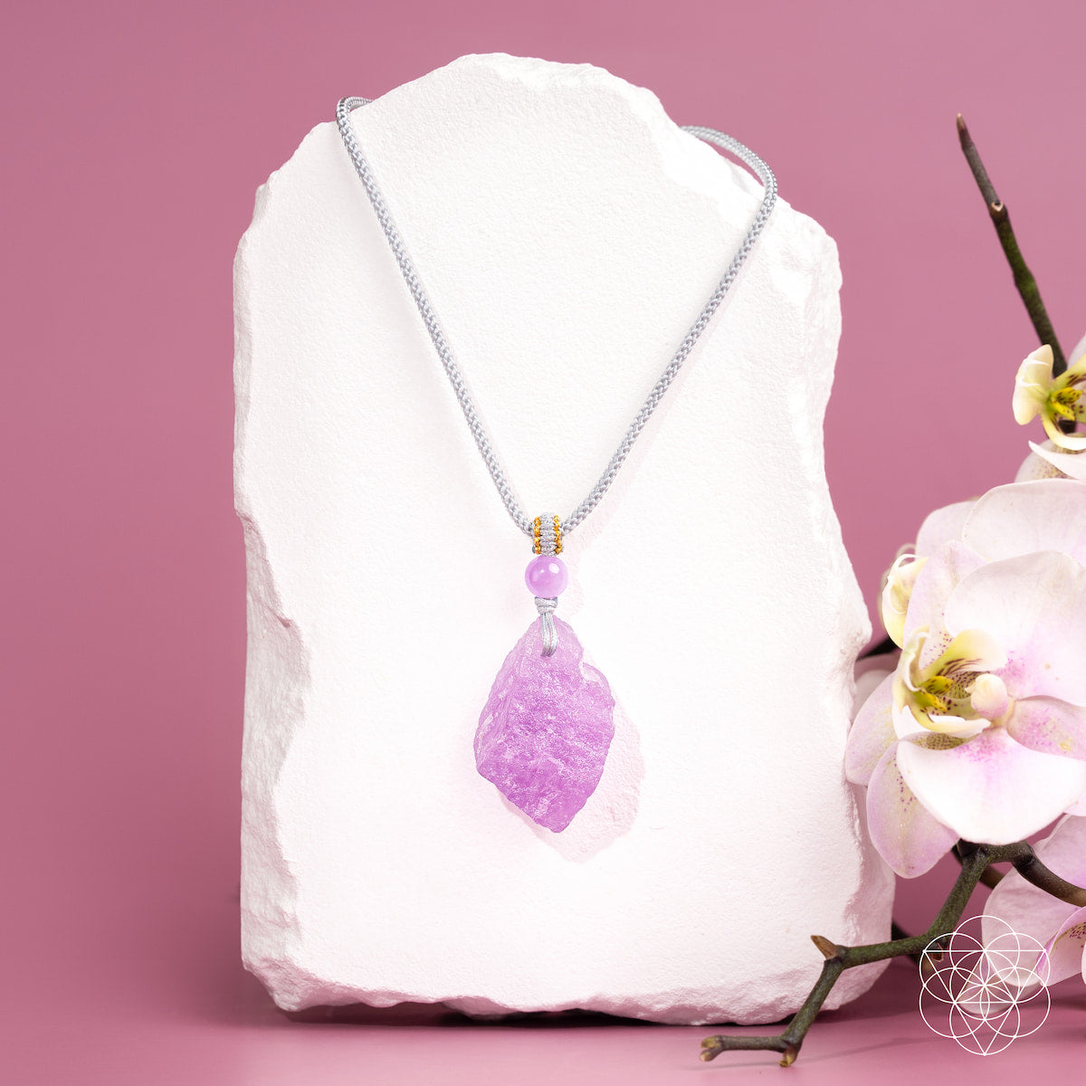 A silver chain necklace with a raw purple crystal pendant displayed on a white stone block against a pink background, with white flowers on the right side.