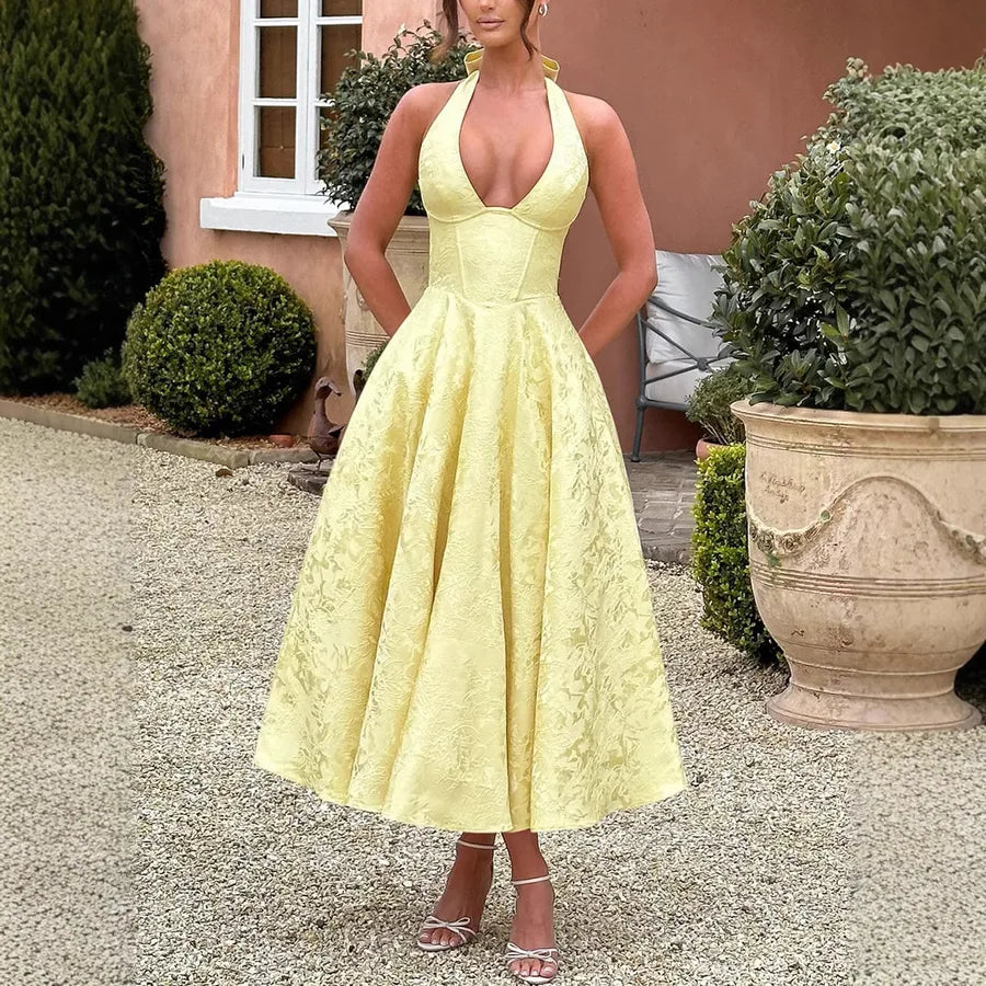 Woman wearing a yellow lace dress standing outdoors with greenery and a building in the background.
