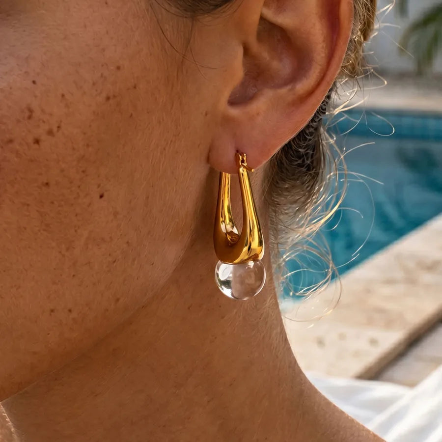 Close-up of a person wearing gold hoop earrings with a clear crystal drop, pool area in the background.