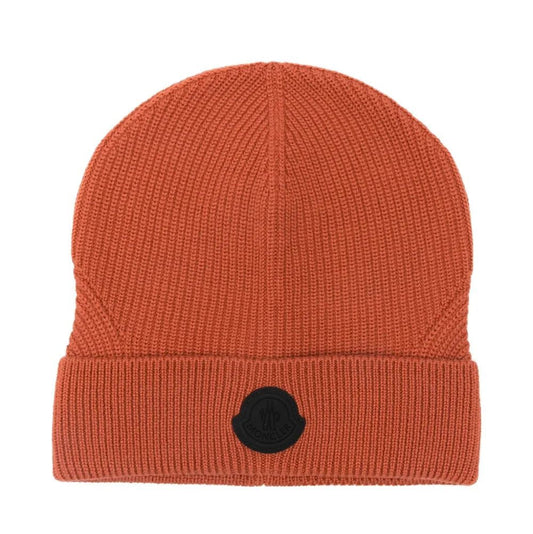 A ribbed knit beanie hat in a warm rust-orange color, featuring a black circular patch on the front of the folded cuff, set against a plain white background.