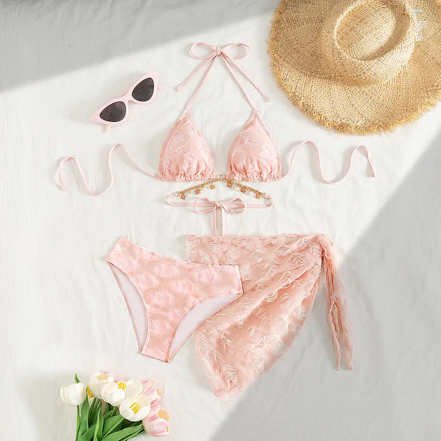 Pink lace bikini set with sunglasses, straw hat, and flowers on a white surface