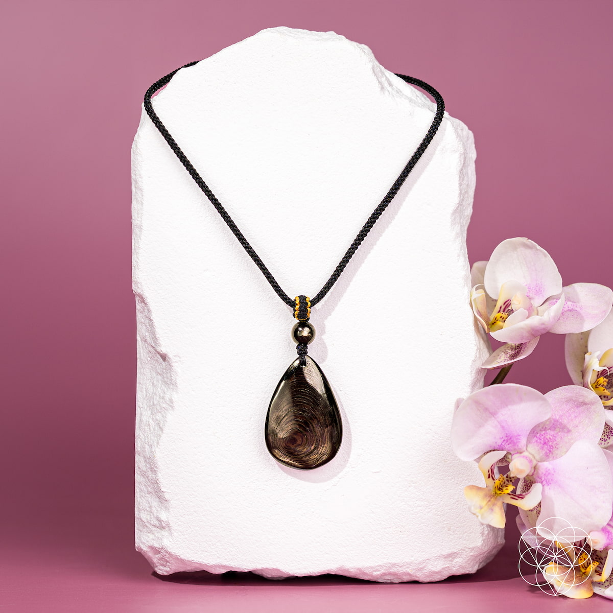 A teardrop-shaped smoky brown gemstone pendant necklace on a black braided cord, displayed on a white stone block with pink orchids on a pink background. The ﻿Hypersthene Millionaire Manifestation Pendant. Rare Egyptian Wealth Stone.
