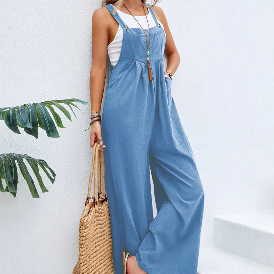 Woman wearing a blue denim jumpsuit with a white top, holding a straw bag against a white wall with green leaves.