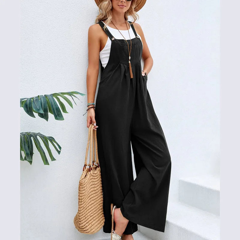 Woman wearing a black jumpsuit with a white top, holding a straw bag against a white wall with green leaf decorations.