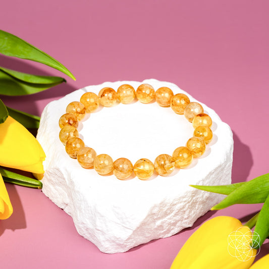 A beaded bracelet made of translucent golden-yellow round beads, displayed on a white stone pedestal with yellow tulips against a pink background.
