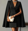Person wearing a black cape dress holding a white clutch against a gray background. Black Blazer Coat Dress