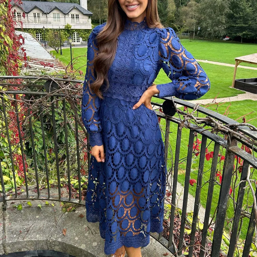 Woman in a blue lace dress standing outdoors with greenery and a building in the background