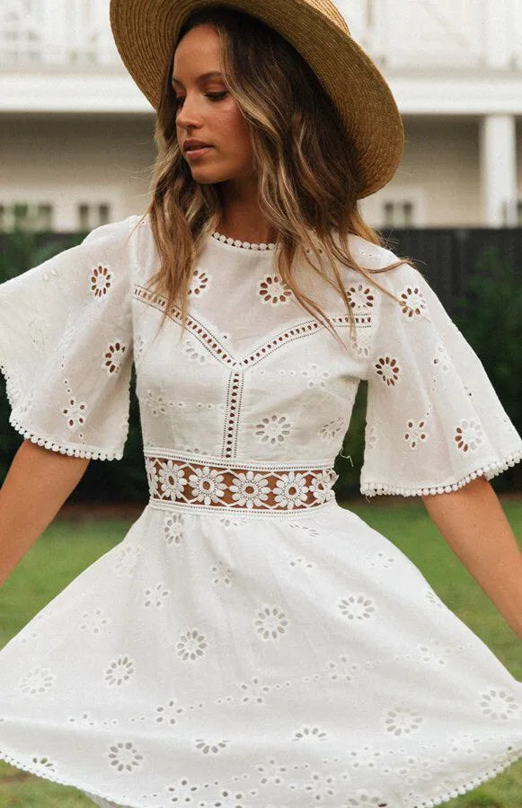 Woman wearing a white dress with floral cut-out details and a straw hat outdoors.
