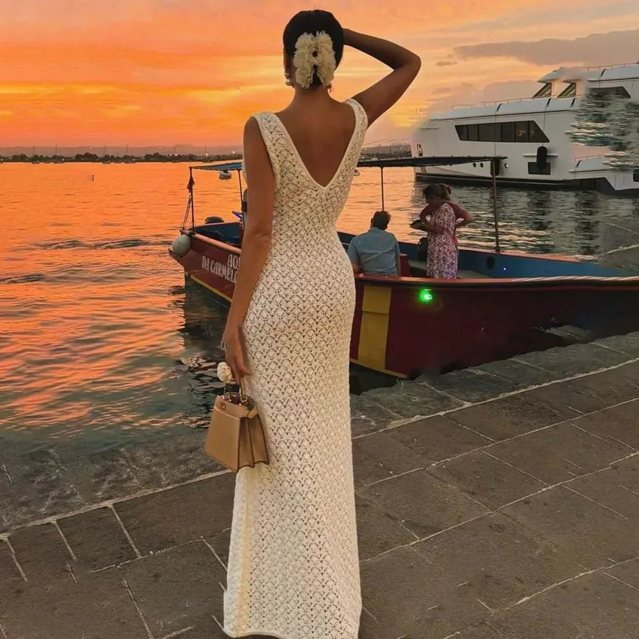 Woman in a cream white dress standing by a waterfront with boats and a sunset. Coastal Breeze Hollow-Out Maxi Sundress – Women's Backless Beach Cover-Up Dress
