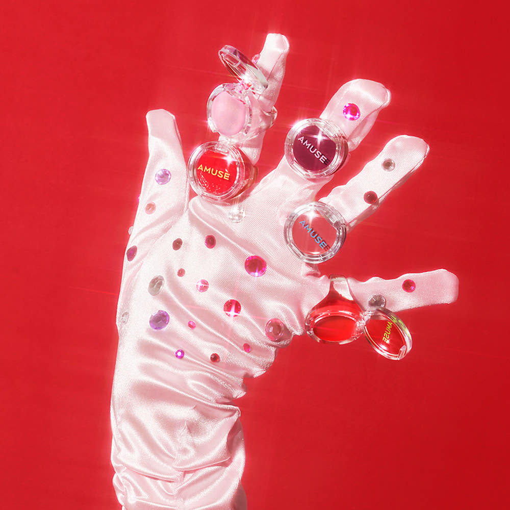 Transparent hand-shaped holder with small bottles labeled 'AMUSE' lip balm rings on a red background. Lip balm finger rings. Red to purple shades.