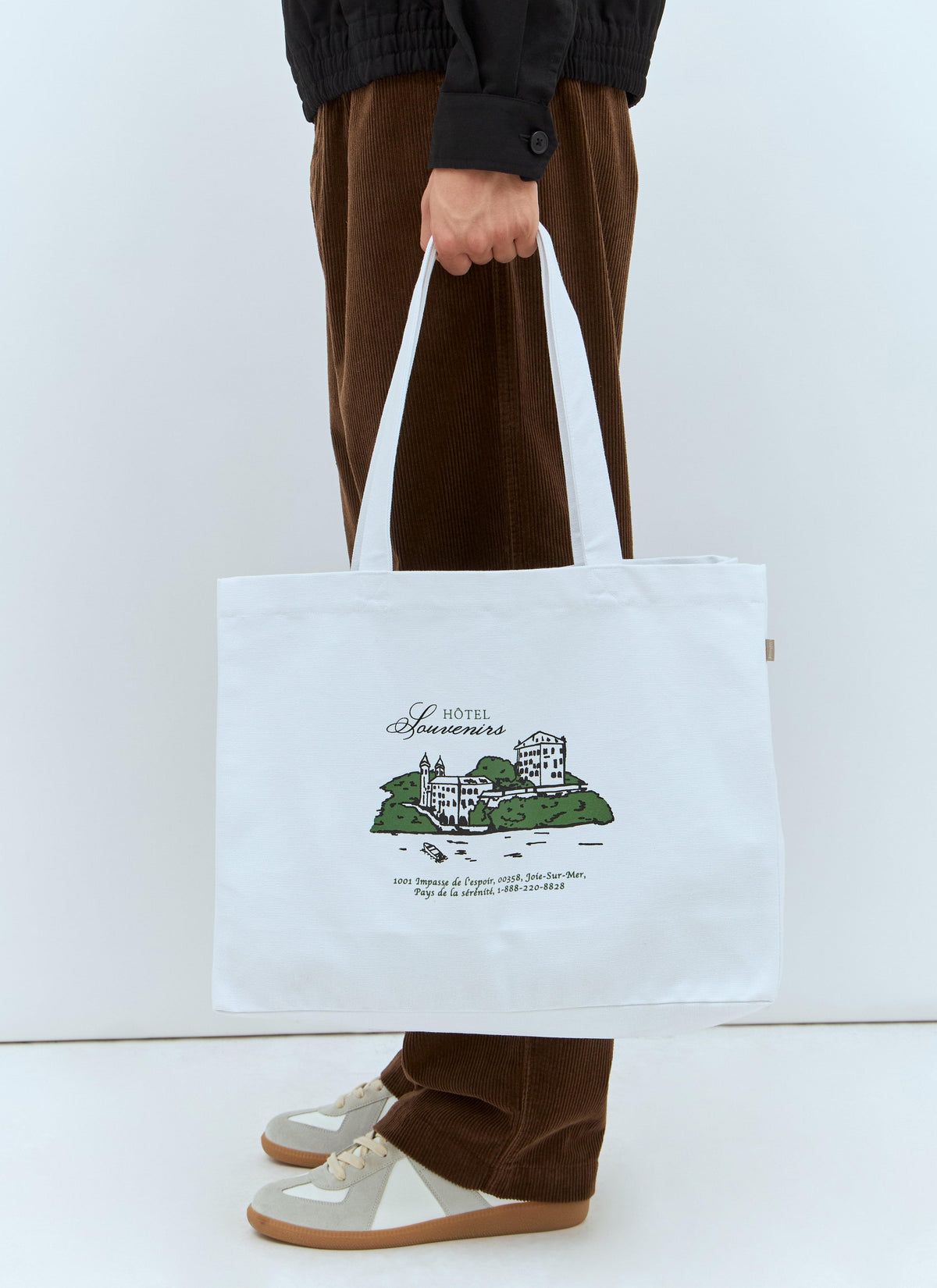 A person wearing brown corduroy pants and beige loafers holds a white canvas tote bag with a black and green illustration of a building and French text. The background is a clean white studio setting.