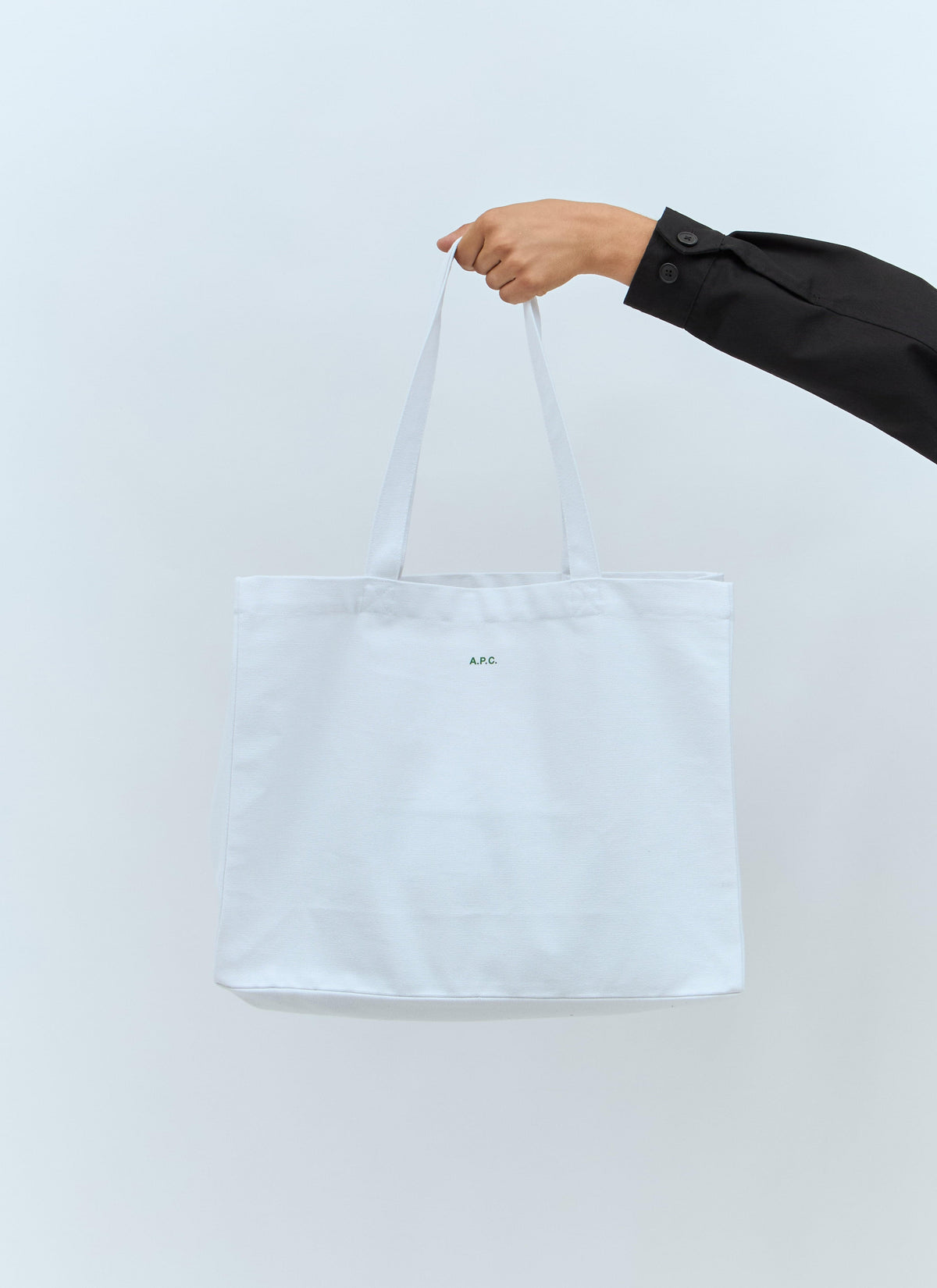 A white canvas tote bag with two handles, held by a person's hand wearing a black sleeve, against a light gray background.