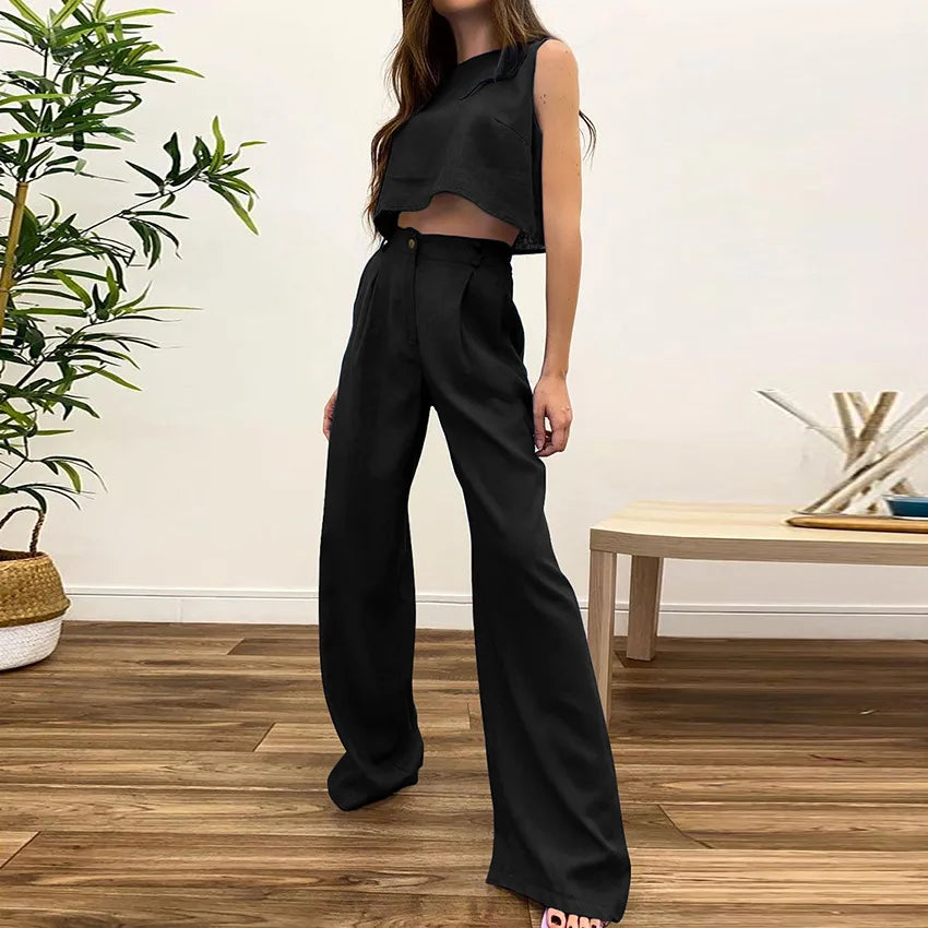 Person wearing a black sleeveless top and wide-leg pants in a room with wooden flooring and a plant.
