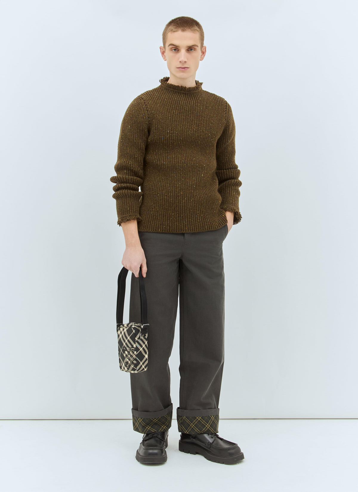 Man wearing a brown sweater and gray pants holding a patterned bag against a white background