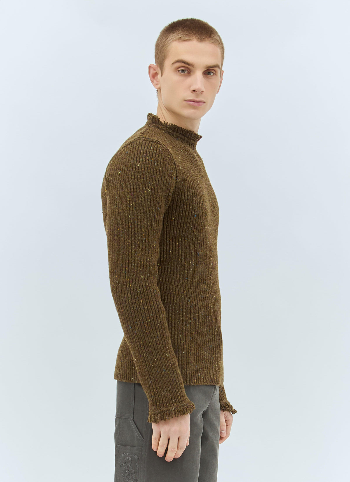 A man wearing a ribbed knit mock neck sweater in a dark brown color with subtle flecks, paired with gray trousers, photographed against a light gray studio background.