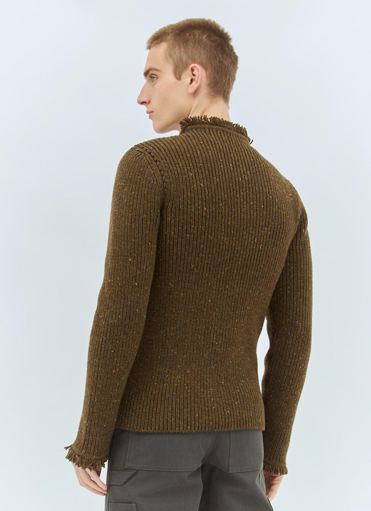 A man wearing a brown ribbed knit sweater with a high mock neck and fringed cuffs, shown from the back against a light blue studio background.