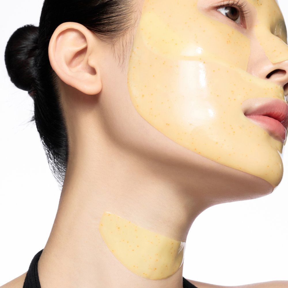 Close-up of a woman's face and neck wearing a smooth, glossy pale yellow skincare mask with small speckles, shown against a plain white background.