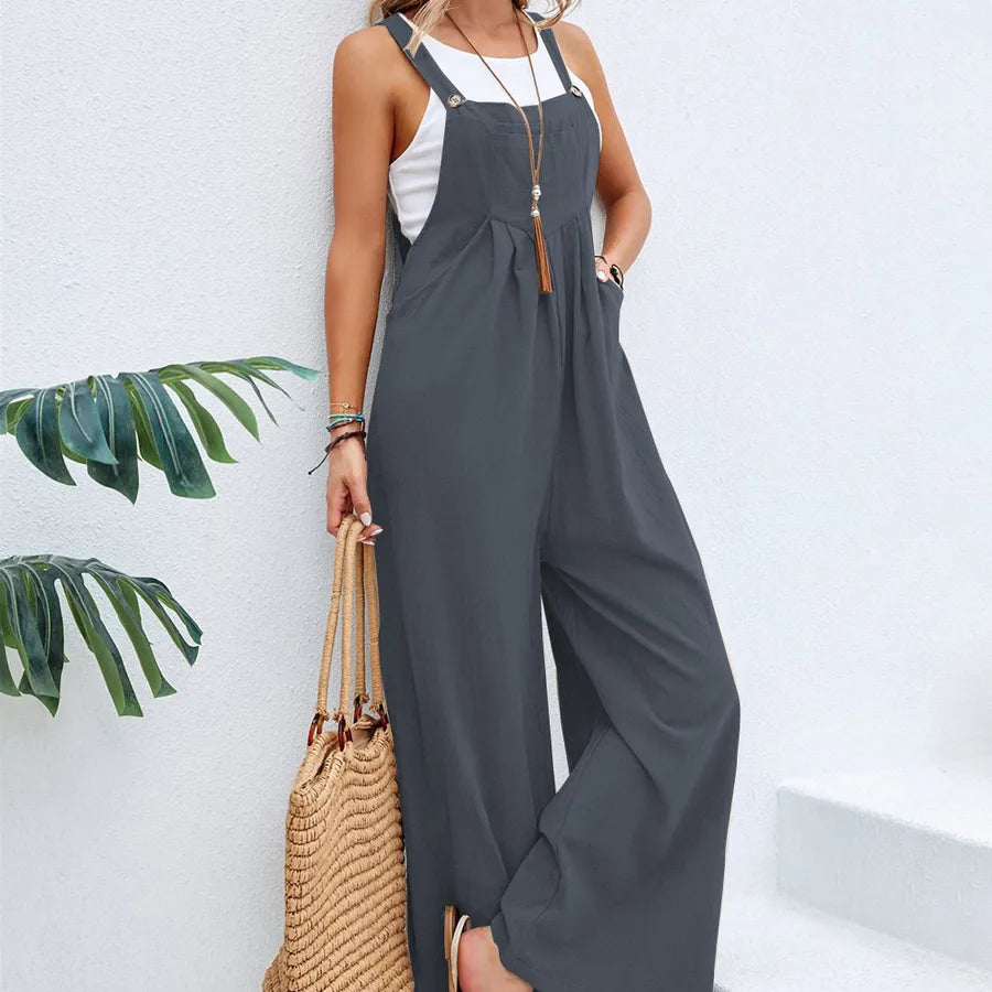 Woman wearing a gray jumpsuit with a white top, holding a straw bag against a white wall with green leaves.