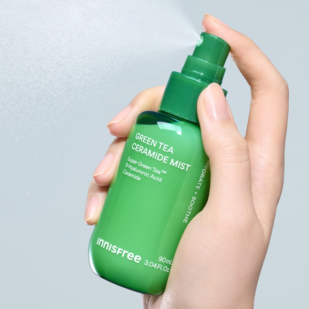 A hand holding an Innisfree Green Tea Ceramide Mist spray bottle, with the green pump spraying a fine mist against a light gray background.