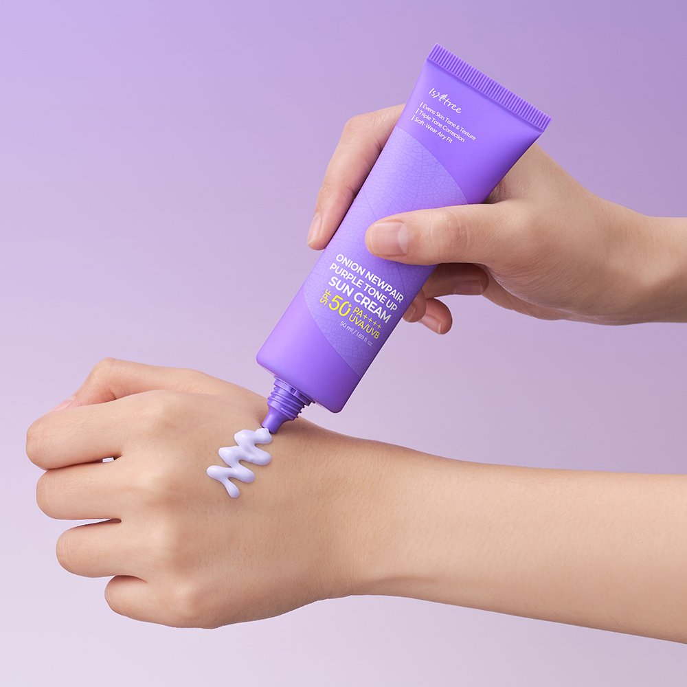 A hand squeezes lavender-colored sunscreen cream from a purple tube onto the back of another hand, set against a solid light purple background. The brand name 'inAra' is visible on the tube.