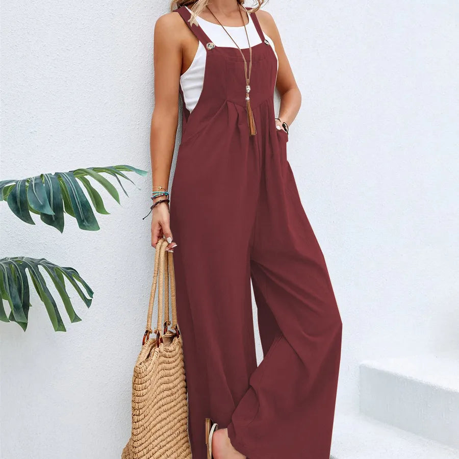 Woman wearing a maroon jumpsuit with a white top, holding a straw bag against a white wall with green leaves.