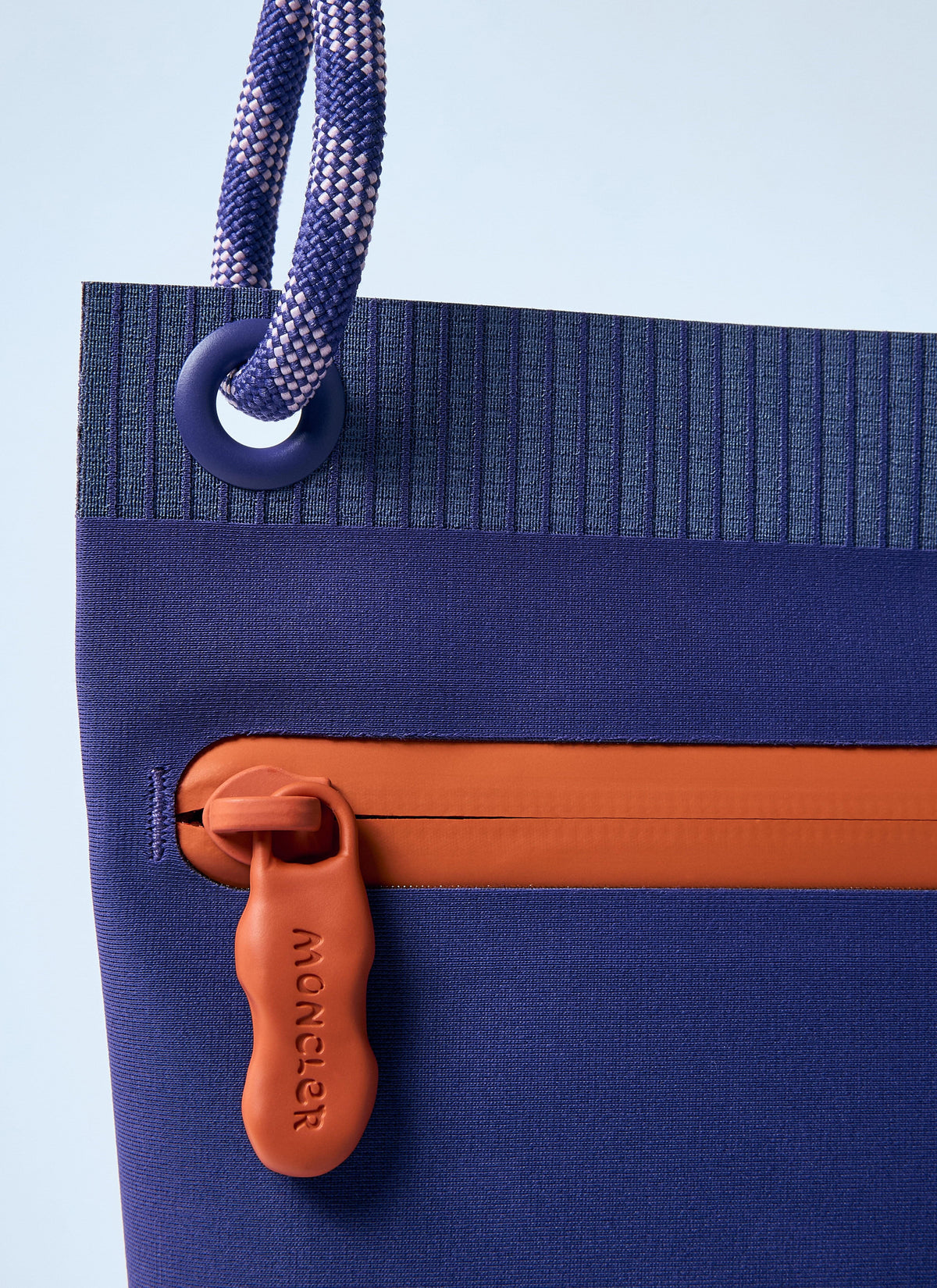 Close-up of a Moncler tote bag in navy blue with a ribbed top panel, a navy-and-white braided rope handle, and an orange zipper pull tab embossed with the Moncler logo.