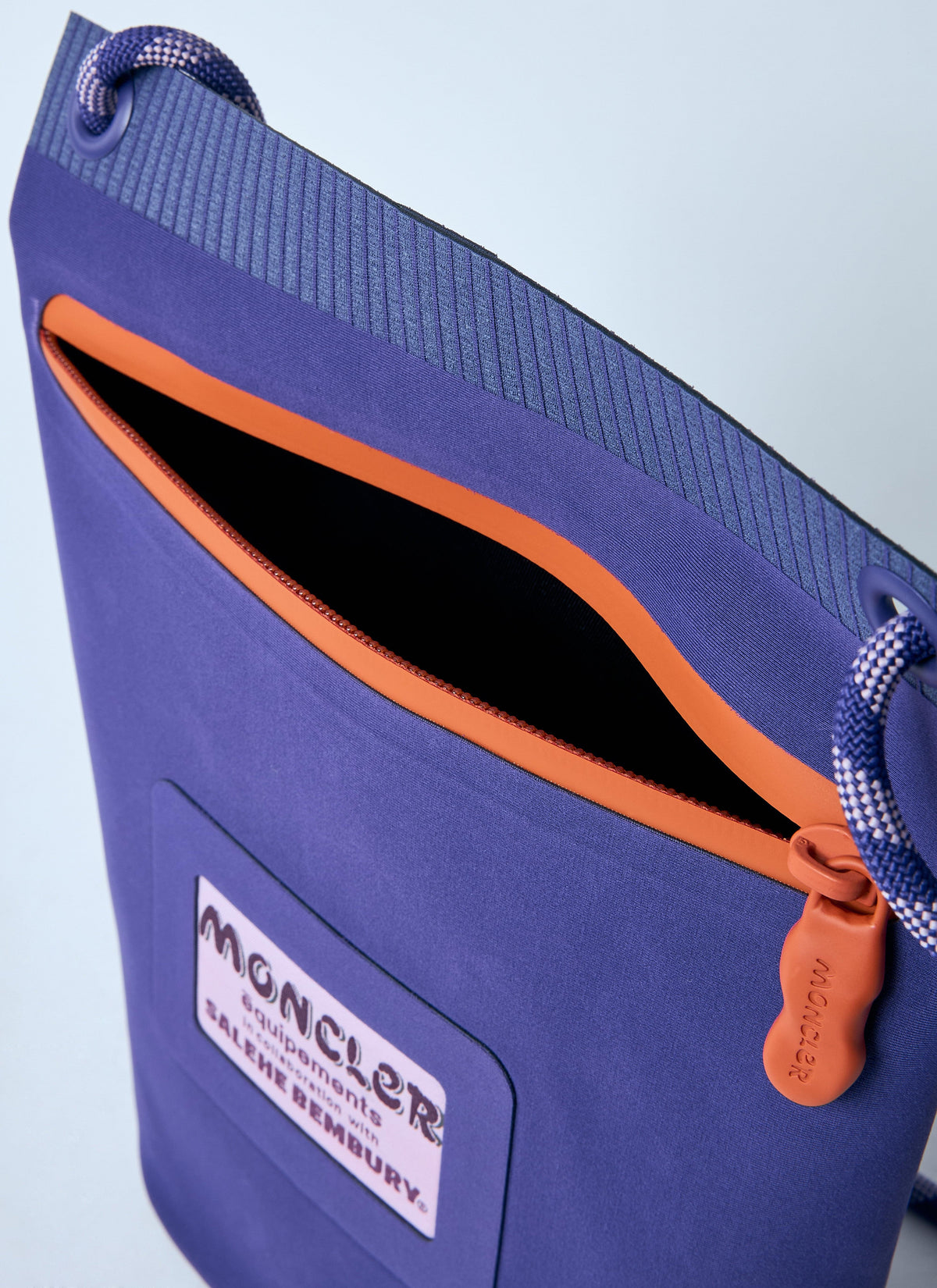Close-up of a purple Moncler drawstring bag with an orange zipper pull and orange trim, featuring a Moncler logo patch on the front.