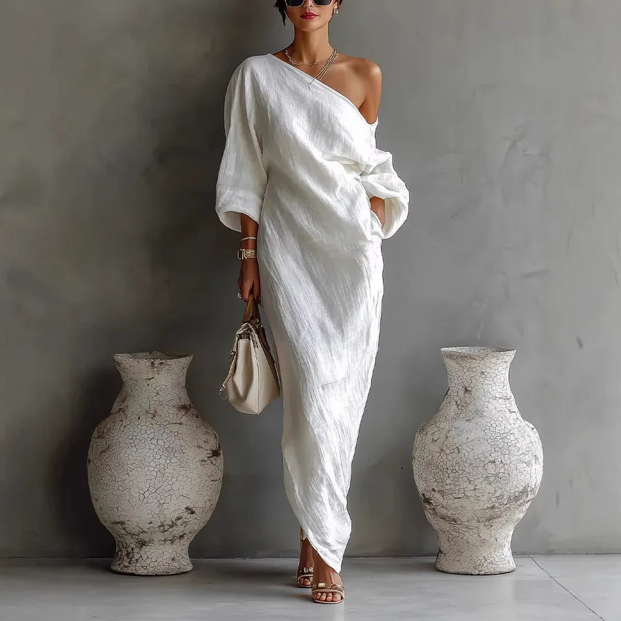 Woman in a white one-shoulder dress standing between two large vases against a gray wall.