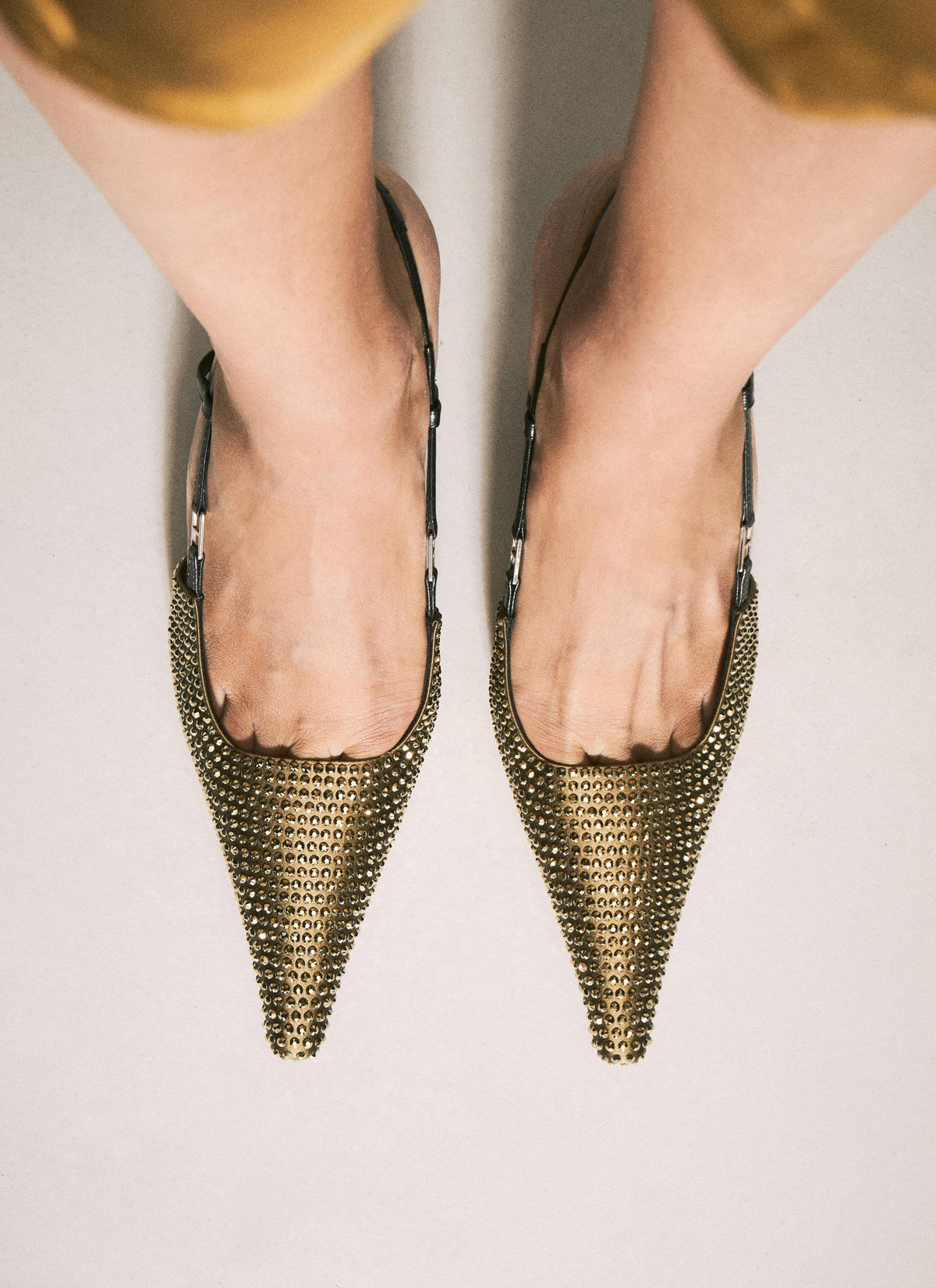 A top-down view of a person wearing pointed slingback heels covered in gold-tone chainmail-style embellishment, shown against a light neutral background.