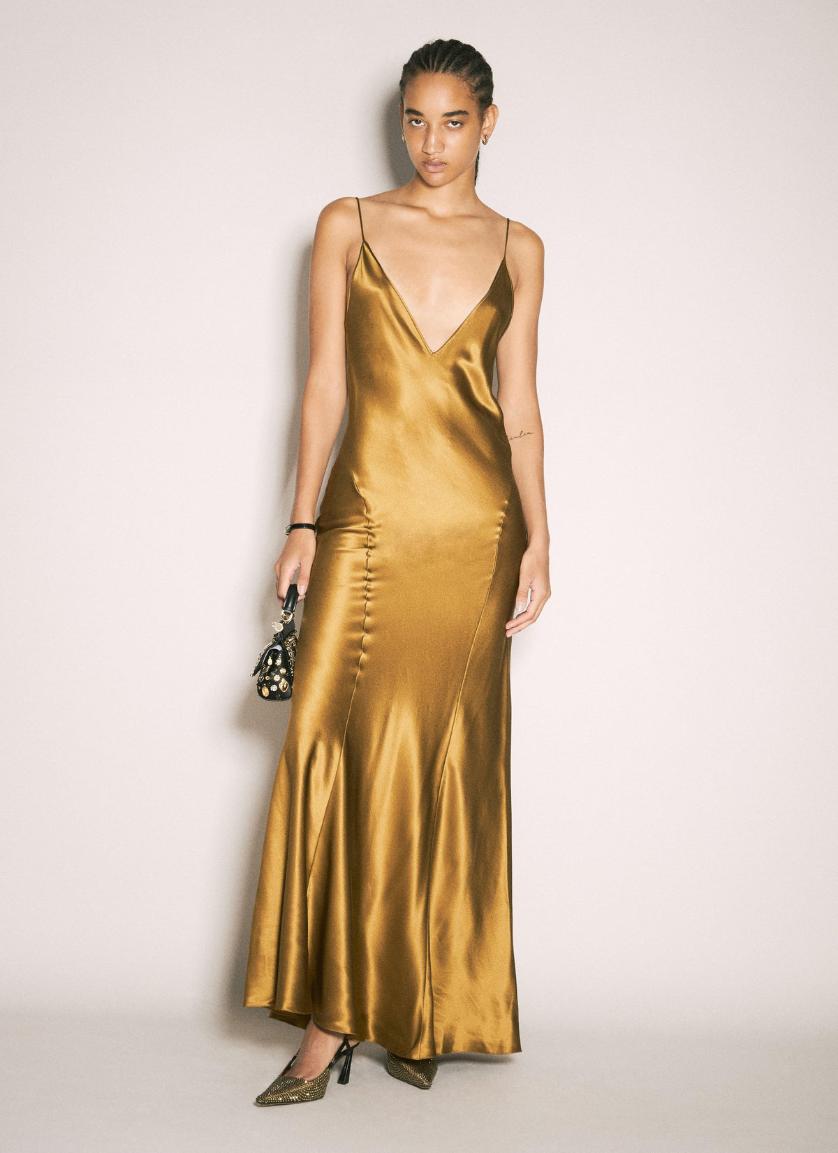 A woman modeling a floor-length satin slip dress in a rich metallic gold color, styled with metallic heels and a small patterned clutch against a plain light background.
