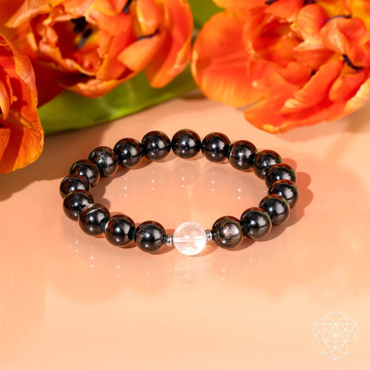 Black beaded bracelet with a clear crystal bead on a peach background with orange flowers. The Hypersthene Millionaire Manifestation Bracelet - Rare Egyptian Wealth Stone